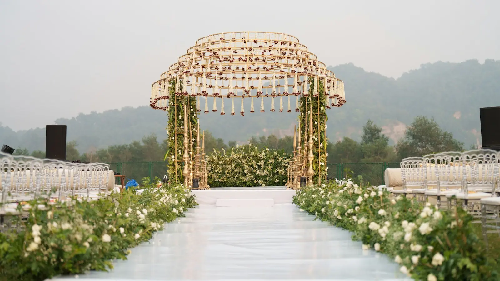 An outdoor wedding mandap of white flowers and greenery at the end of a flower-lined aisle